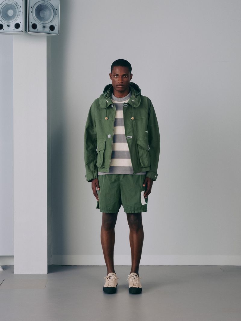 An African American man stands in a studio in a green hooded jacket, striped shirt, green shorts, and sneakers looking at the camera.