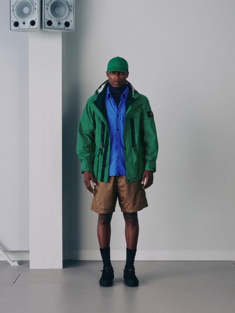 A man in a green jacket, blue shirt, and tan shorts poses indoors with speakers in the background, showcasing casual and stylish attire.