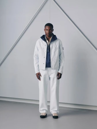 Black man standing in a light blue jacket and white pants against a geometric studio backdrop for Stone Island Lookbook SS26.