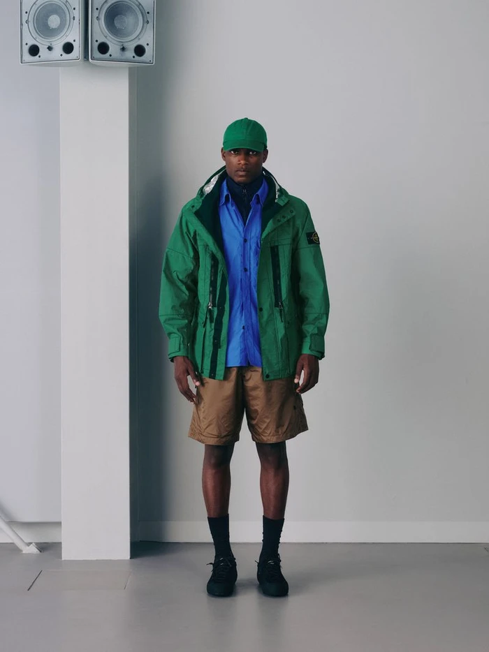 Akin Muyiwa wearing a green technical jacket, blue shirt, tan shorts, and green cap for Stone Island SS26.