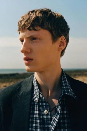 Portrait of a young male model with brown hair wearing a plaid shirt and dark jacket outdoors against the ocean