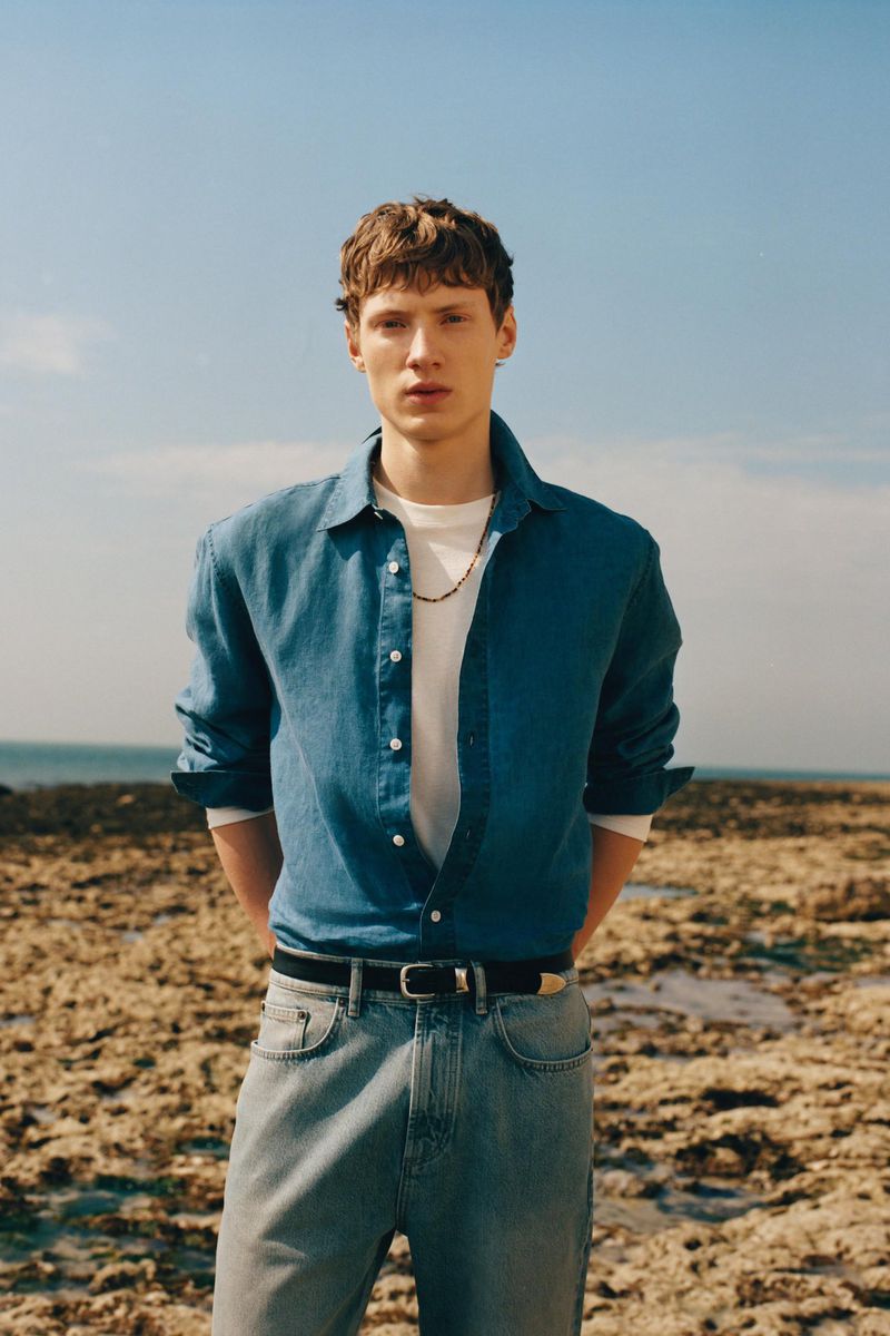 Model Braien Vaiksaar wearing an unbuttoned blue shirt, white t-shirt, and denim jeans on a rocky coastline for Zara Origins.