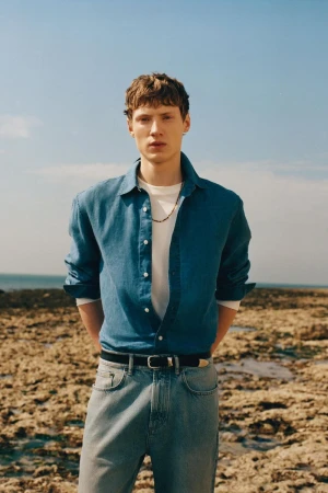 Model Braien Vaiksaar wearing an unbuttoned blue shirt, white t-shirt, and denim jeans on a rocky coastline for Zara Origins.