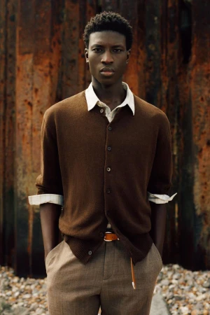 Cheikh Amar Diaw in a brown knit cardigan, white collared shirt, and beige trousers against a rusted metal backdrop.