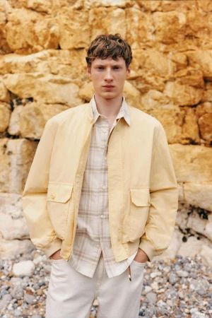 Model Braien Vaiksaar standing against a rugged stone rock face wearing a pale yellow bomber jacket and white trousers.