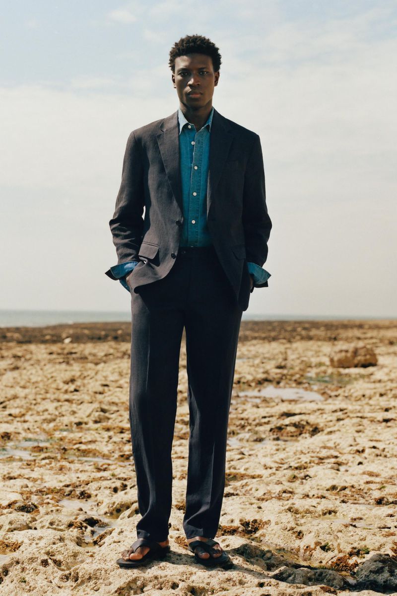 A Black man standing on a rocky coast wearing a dark tailored suit, blue button-down shirt, and sandals for Zara Origins.