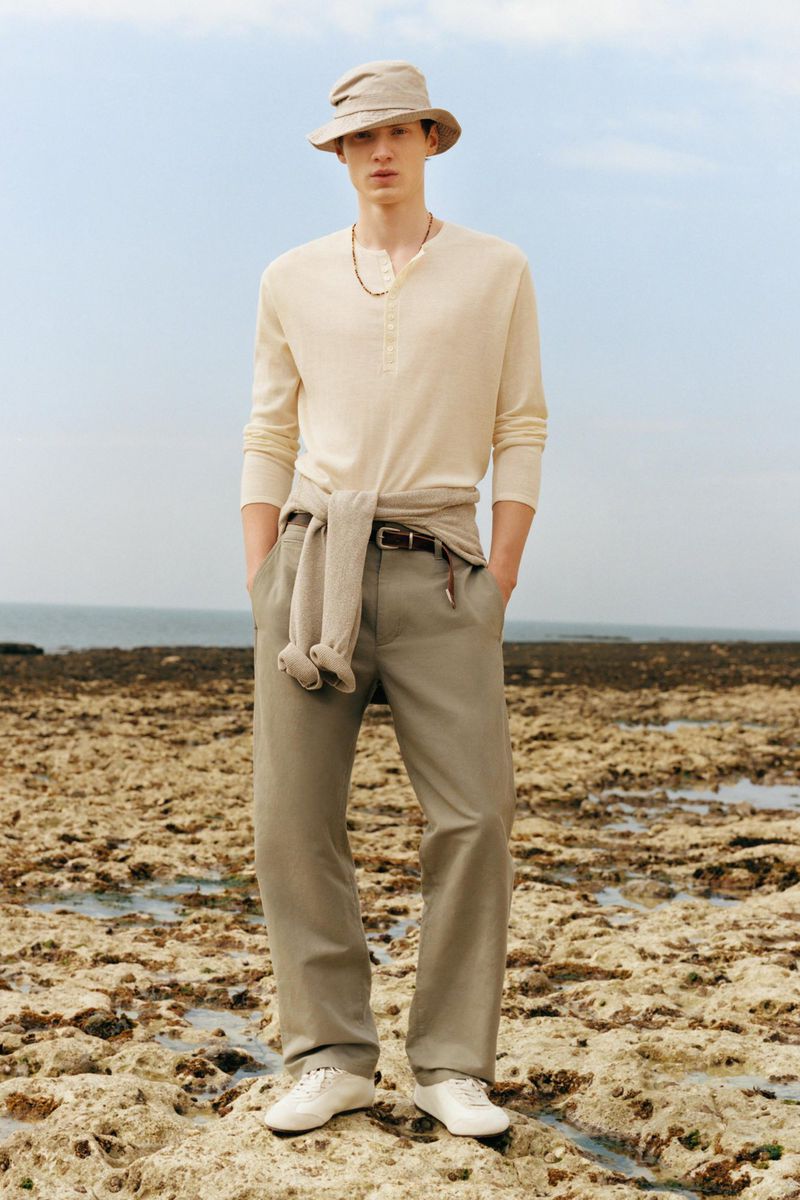 Model Braien Vaiksaar in a cream henley shirt, khaki trousers, and a bucket hat standing on a rocky shoreline.