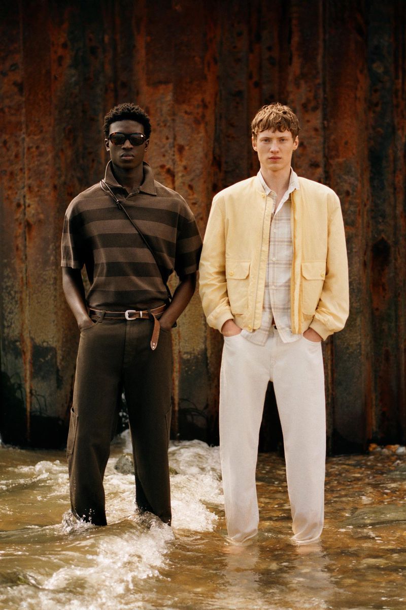Two male models stand in shallow seawater before a rusted corrugated wall wearing a striped polo and a pale yellow jacket.