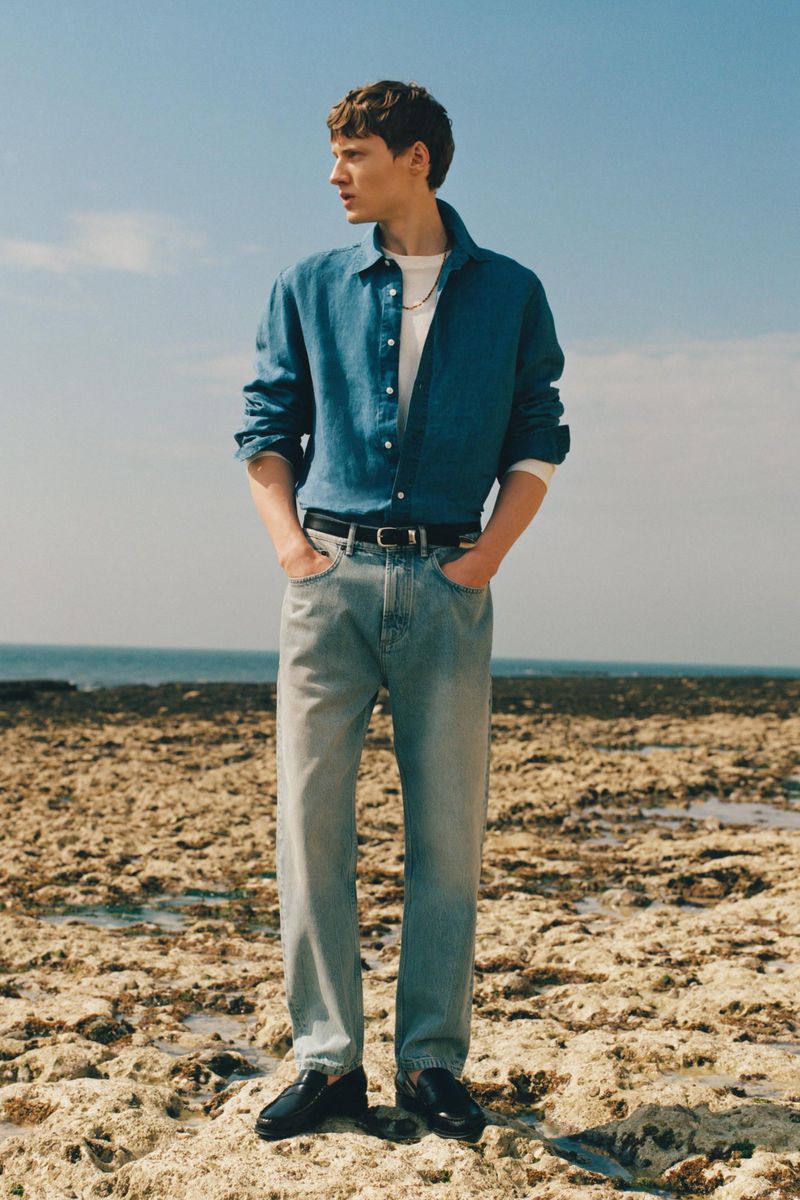 Male model wearing a blue denim shirt and light wash jeans standing on a rocky shore overlooking the ocean.