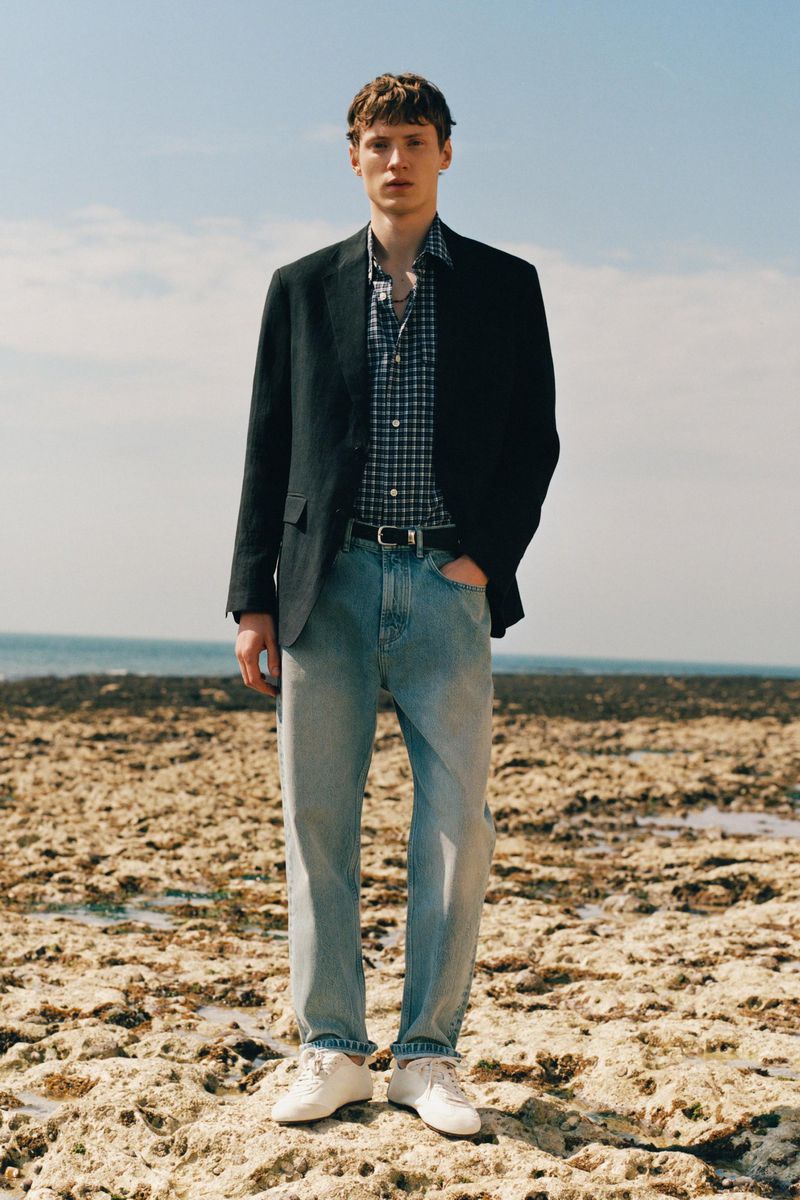 Model Braien Vaiksaar wearing a dark blazer, gingham shirt, and light wash denim jeans standing on a rocky shoreline.