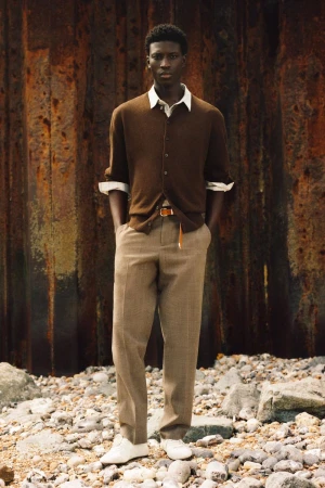 Model Cheikh Amar Diaw wearing a brown knit cardigan and beige trousers standing on pebbles against a rusted metal backdrop.