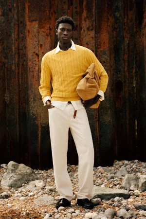 Model Cheikh Amar Diaw stands in a yellow cable-knit sweater and cream trousers against a weathered metal wall on a pebble beach.