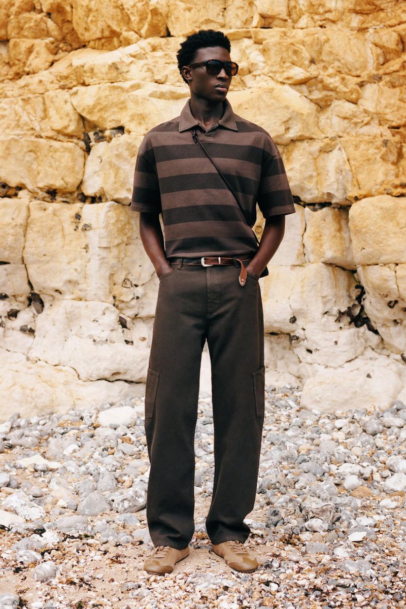 Model standing against a rocky cliff wearing a brown striped polo shirt, wide-leg dark brown trousers, and dark sunglasses.