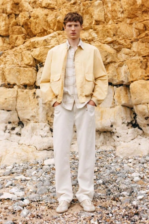 Model standing outdoors wearing a light yellow jacket, patterned shirt, and white trousers against a rocky cliff face
