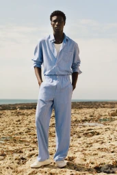Model Cheikh Amar Diaw wearing a light blue striped shirt and matching trousers on a rocky coastline for the Zara Origins campaign.