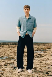 Model Braien Vaiksaar standing on a rocky coast wearing a light blue short-sleeve shirt over a long-sleeve layer and dark trousers.