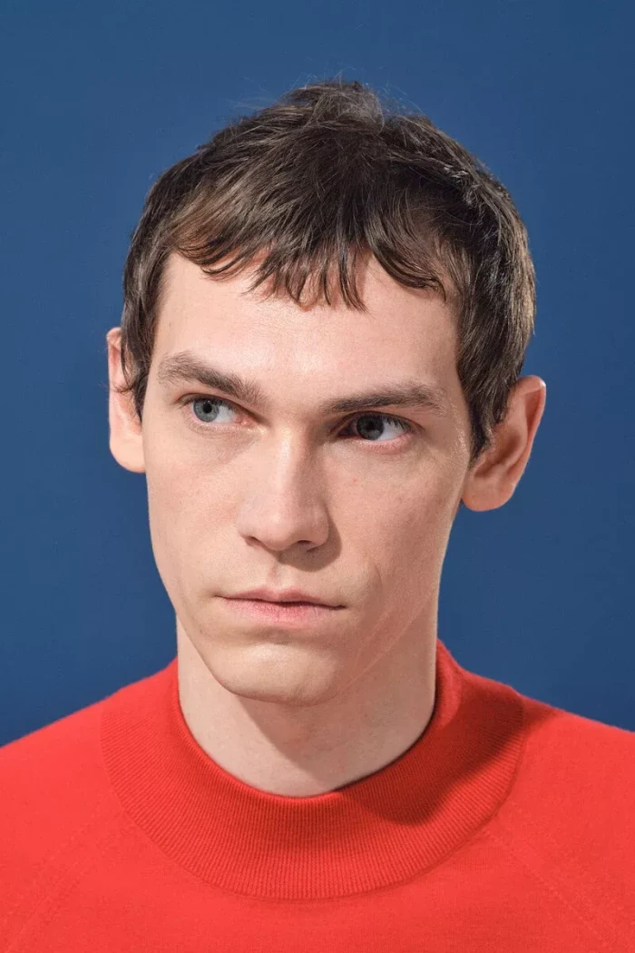 Model Aleksandr Gordeev wearing a vibrant red crewneck top against a solid blue backdrop.