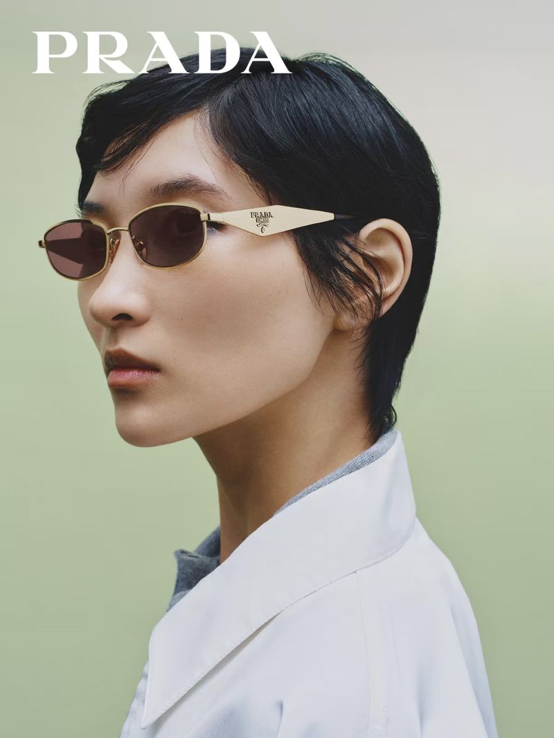 East Asian model with short dark hair wearing gold-toned Prada sunglasses and a white collared shirt against a soft green backdrop.