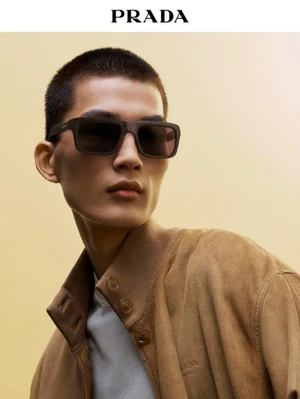 East Asian male model with buzz cut wearing dark rectangular sunglasses and a tan suede jacket against a warm beige background.
