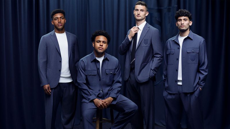 Four men in navy blue BOSS tailored suits and utility jackets standing and sitting against a dark curtain background.