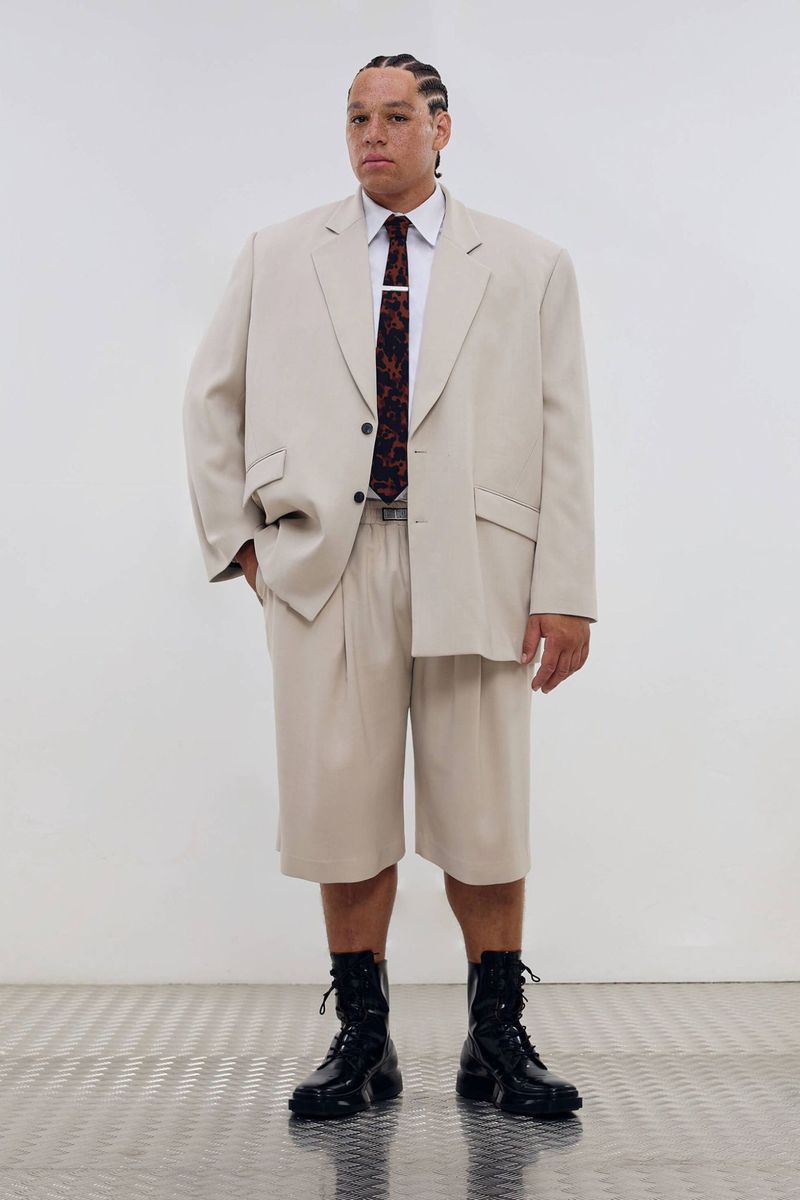 Man in an oversized beige suit with a black patterned tie and black lace-up boots stands in a white interior.