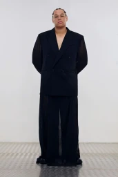 Model wearing an oversized black double-breasted blazer and sheer black trousers against a white studio wall with metal tread flooring.