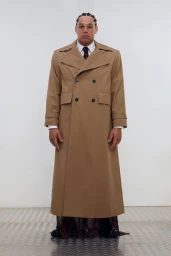 Model in a full-length camel double-breasted trench coat standing on diamond plate flooring against a white backdrop.