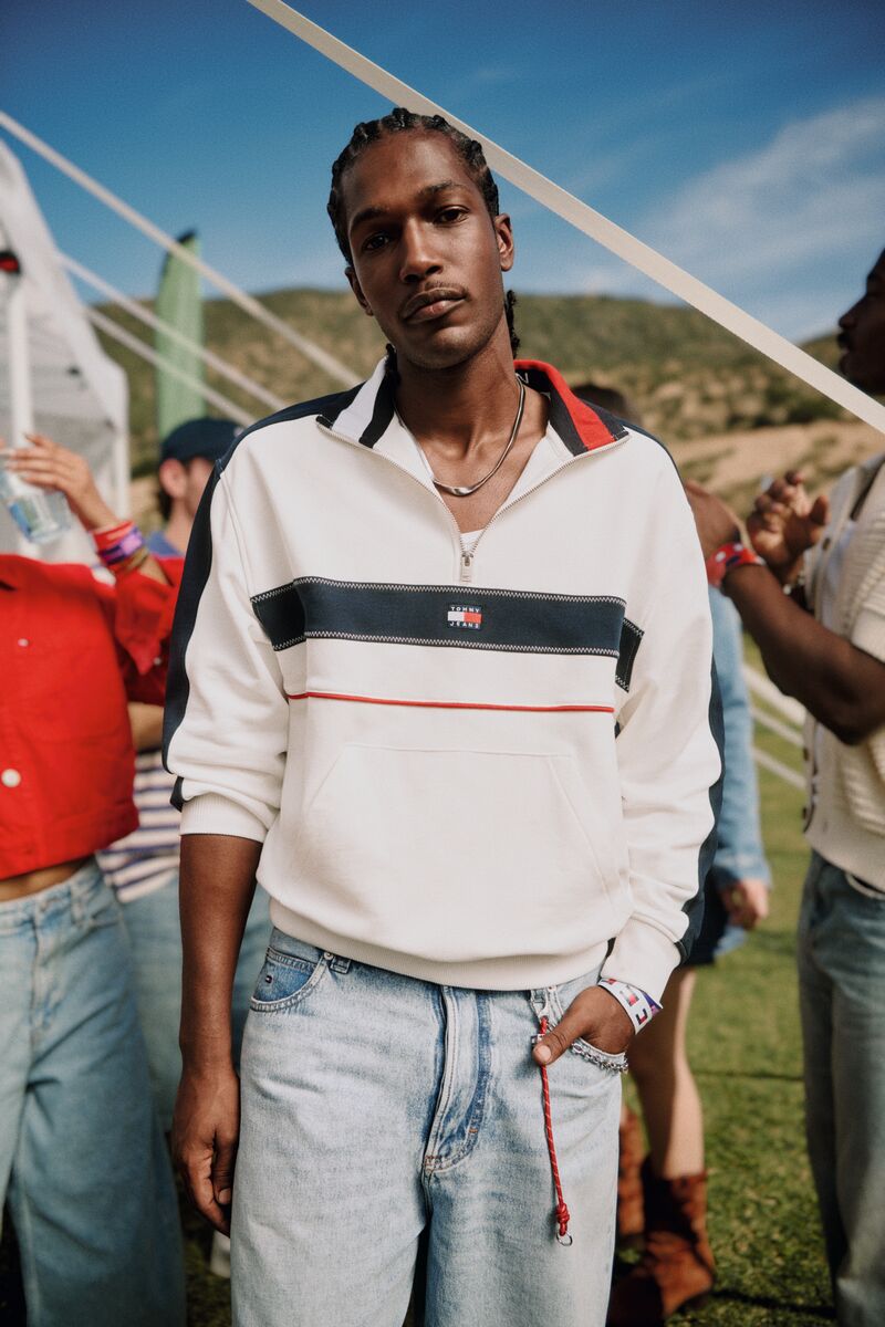 A Black man wearing a white quarter-zip sweatshirt with stripe details and light-wash jeans stands outdoors with people on a lawn.