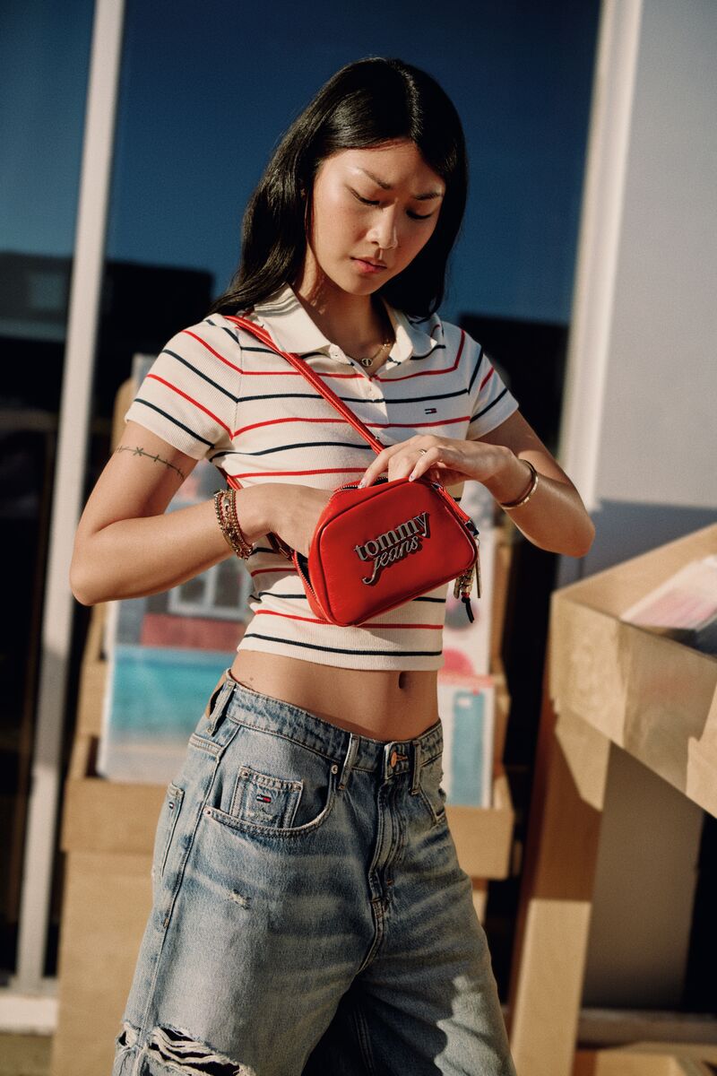 Model wearing a striped crop polo and distressed jeans reaches into a red Tommy Jeans crossbody bag in direct sunlight.