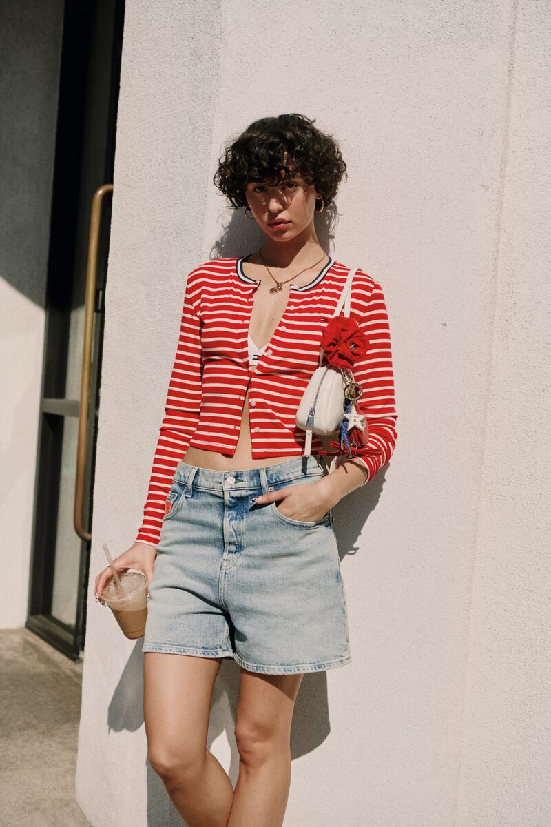 A model with short curly hair leaning against a sunlit exterior wall wearing a red and white striped cardigan and denim shorts.