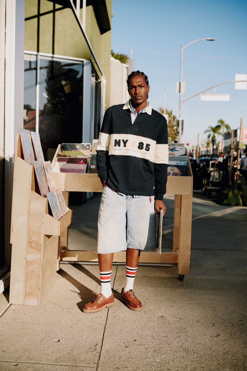 A Black man standing on a sidewalk next to a record bin wearing a navy polo shirt, light denim shorts, and brown shoes.