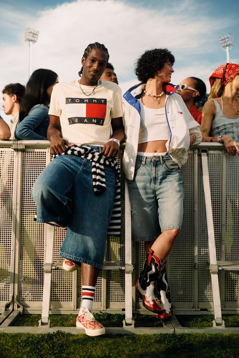 Two models leaning against metal barriers outdoors wearing Tommy Jeans branded apparel and denim shorts