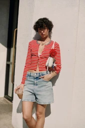 A model with short curly hair leaning against a sunlit exterior wall wearing a red and white striped cardigan and denim shorts.