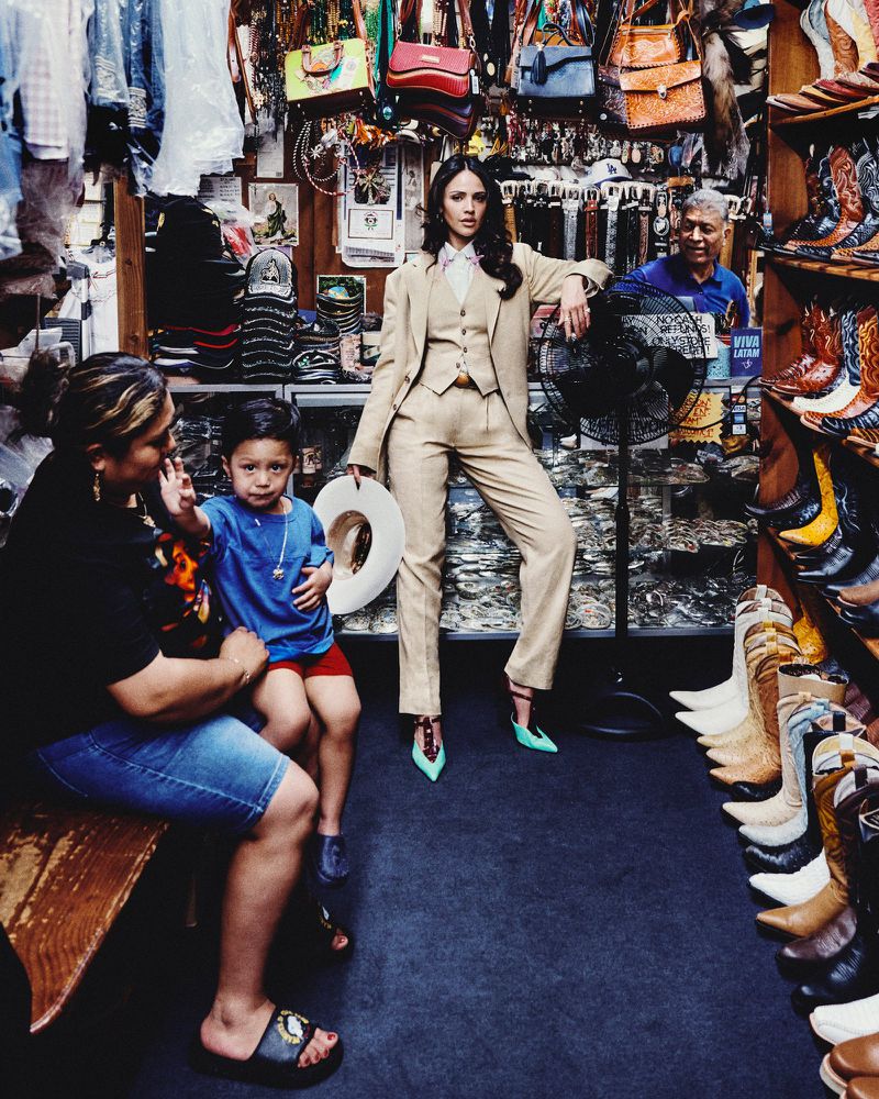 Eiza Gonzalez posing in a tailored beige three-piece suit holding a white hat inside a Western boot and leather goods shop.
