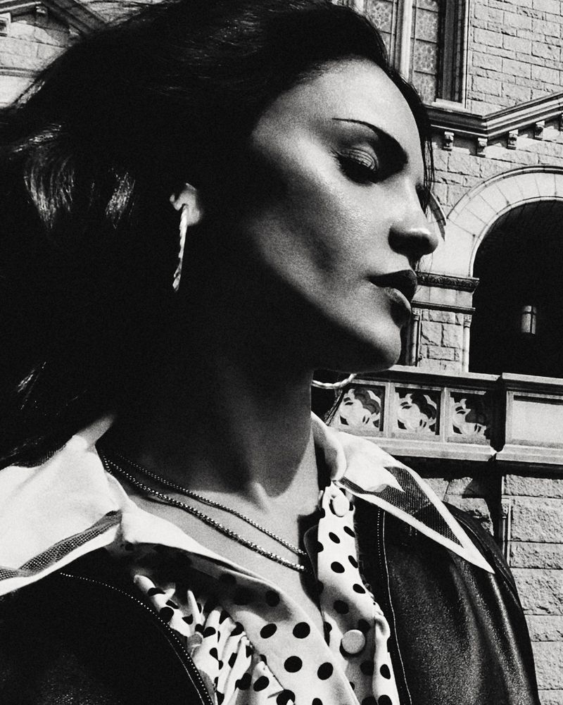 Black and white profile portrait of Eiza Gonzalez wearing a polka dot collar shirt and leather jacket beside a stone building.