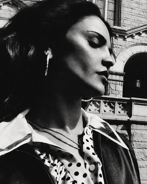 Black and white profile portrait of Eiza Gonzalez wearing a polka dot collar shirt and leather jacket beside a stone building.