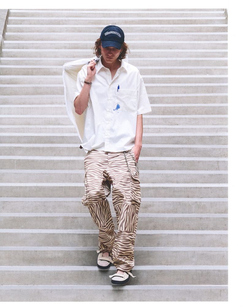 Young man descends concrete steps wearing baseball cap, patterned pants, and a white shirt with a white jacket over his shoulder.