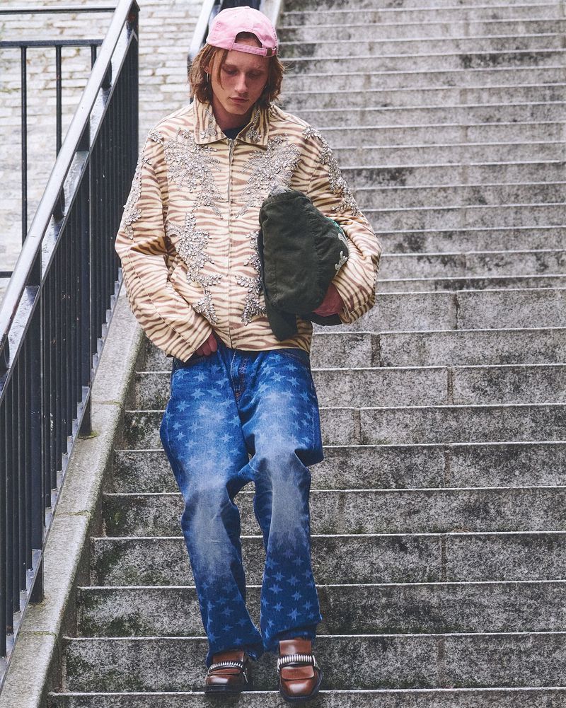 Adult male with long hair descends concrete stairs outdoors wearing jeans with star pattern, and printed tiger-stripe jacket.