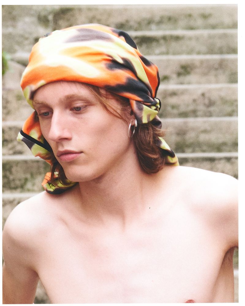 Portrait of a bare-chested young man wearing a colorful headscarf, gazing at the camera with a serene expression and neutral background.