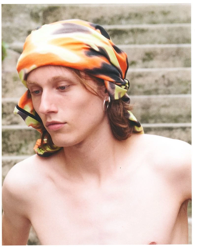 Shirtless young man wearing an orange patterned headscarf and silver hoop earring outdoors on stone steps.