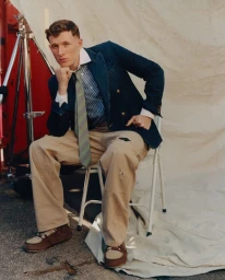 Young man in a navy blazer, striped tie, and beige trousers seated on a white folding chair before a canvas backdrop.