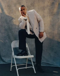 Man in a light double-breasted coat and dark trousers posing with one foot on a white folding chair before a textured grey backdrop.