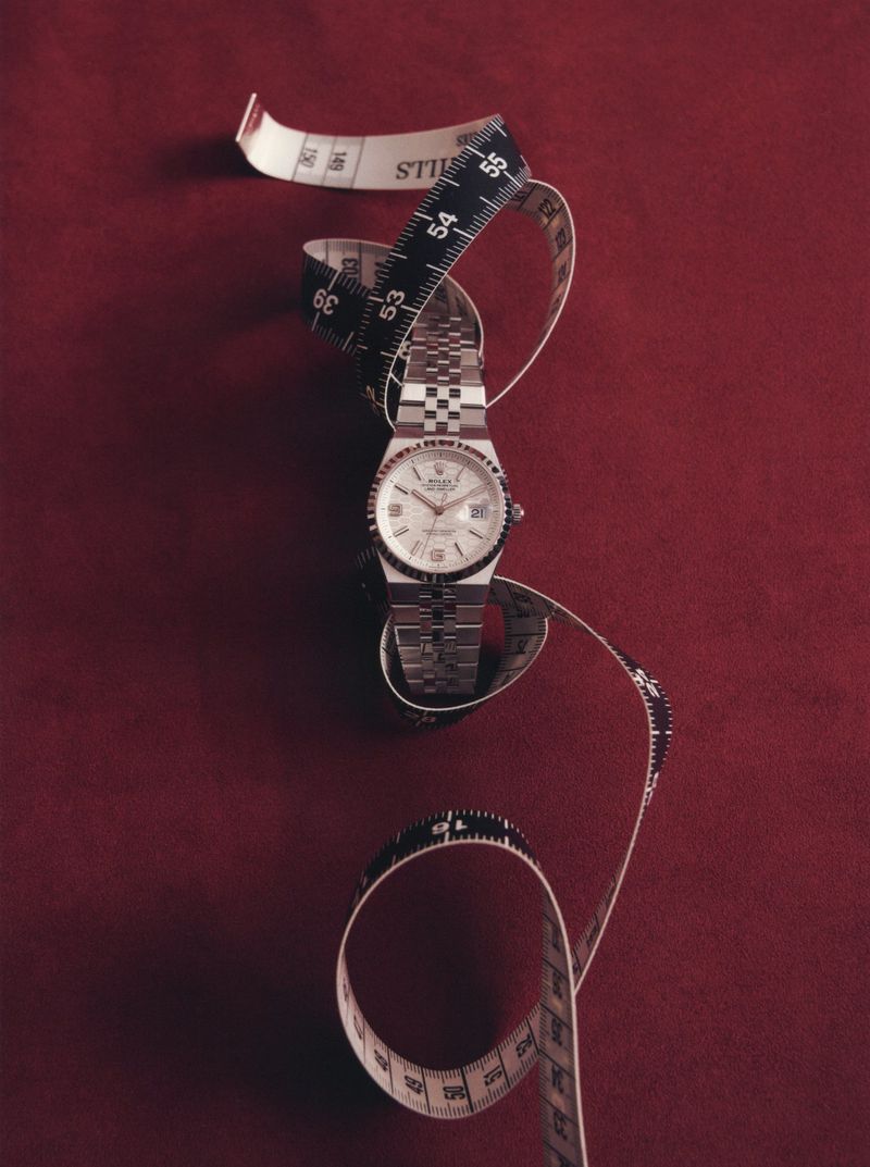 Silver stainless steel wristwatch surrounded by a curled black and white tailor measuring tape on a burgundy fabric surface.