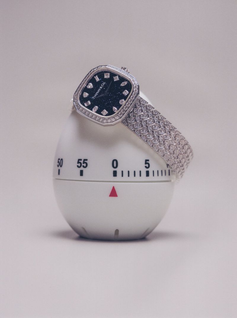 Diamond-encrusted luxury watch draped over a white egg-shaped kitchen timer on a pale neutral seamless backdrop.