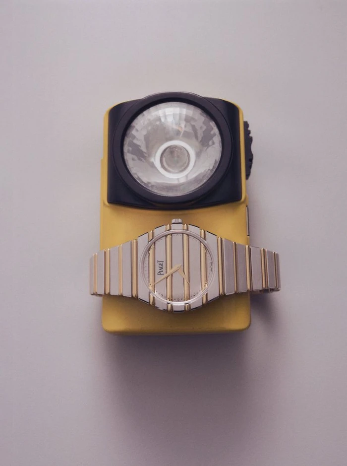 Two-tone gold and silver wristwatch resting across a yellow and black utility flashlight on a pale neutral backdrop.