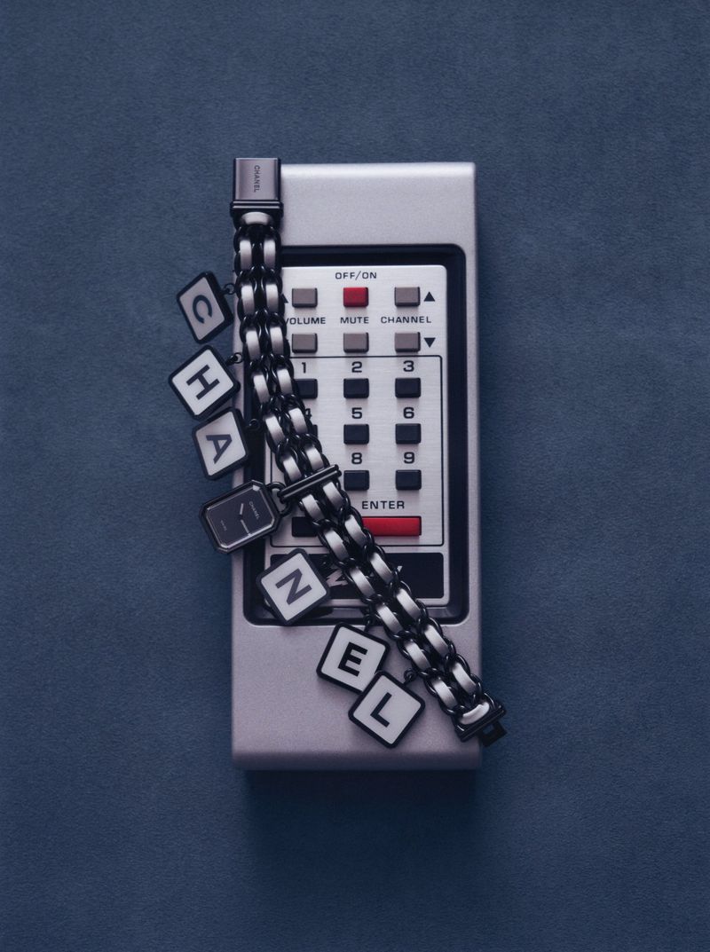 Chanel letter charm watch draped across a vintage grey remote control against a dark blue background.