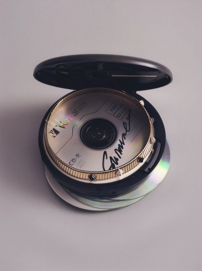 An open black portable CD player resting on a stack of CDs, containing a Verbatim CD-R bordered by a studded gold watch bezel.