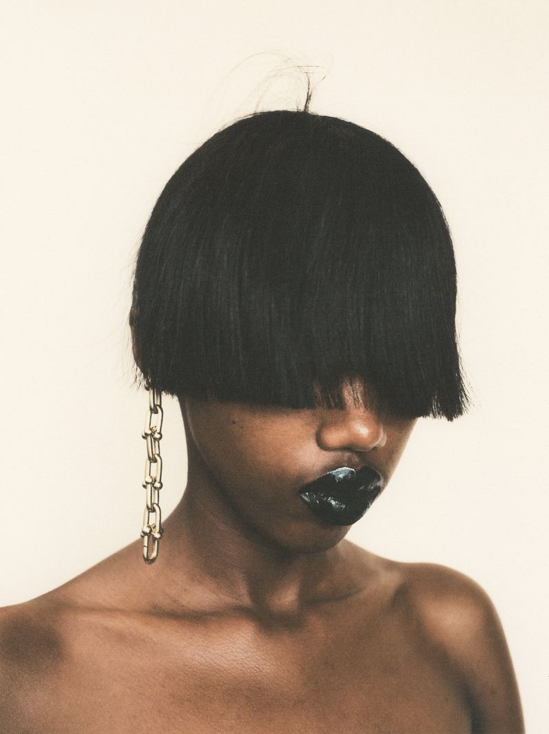 Esther Niyirera with a blunt black bob covering her eyes, glossy dark lipstick, and a chain earring against a plain background.