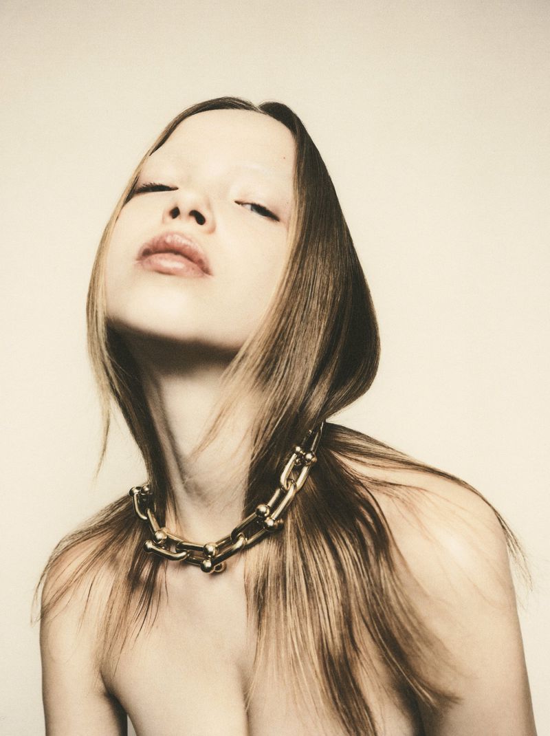 Close-up portrait of a woman with light brown hair tilted back, wearing a chunky gold link chain necklace against a pale backdrop.