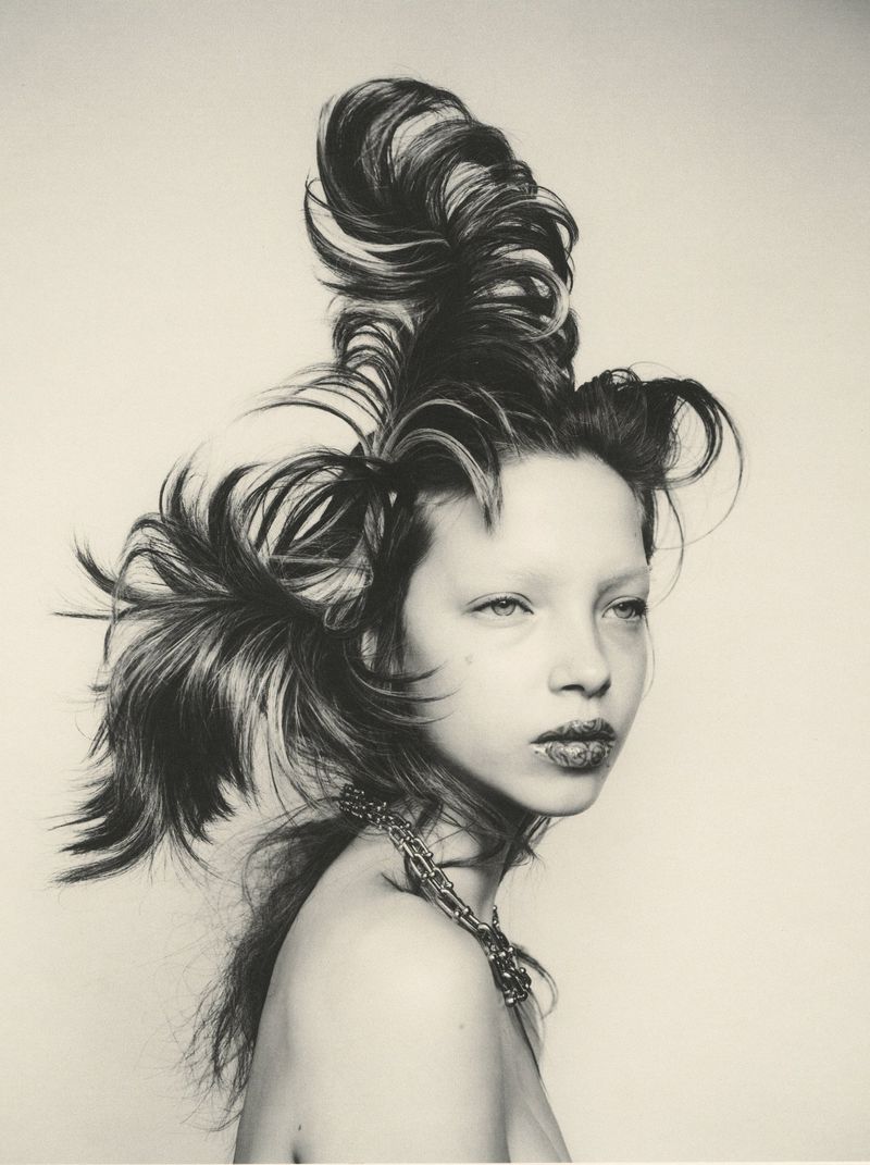 Monochrome editorial portrait of a model with dramatic sculptural swept-up hair wearing a chain necklace against a light backdrop.